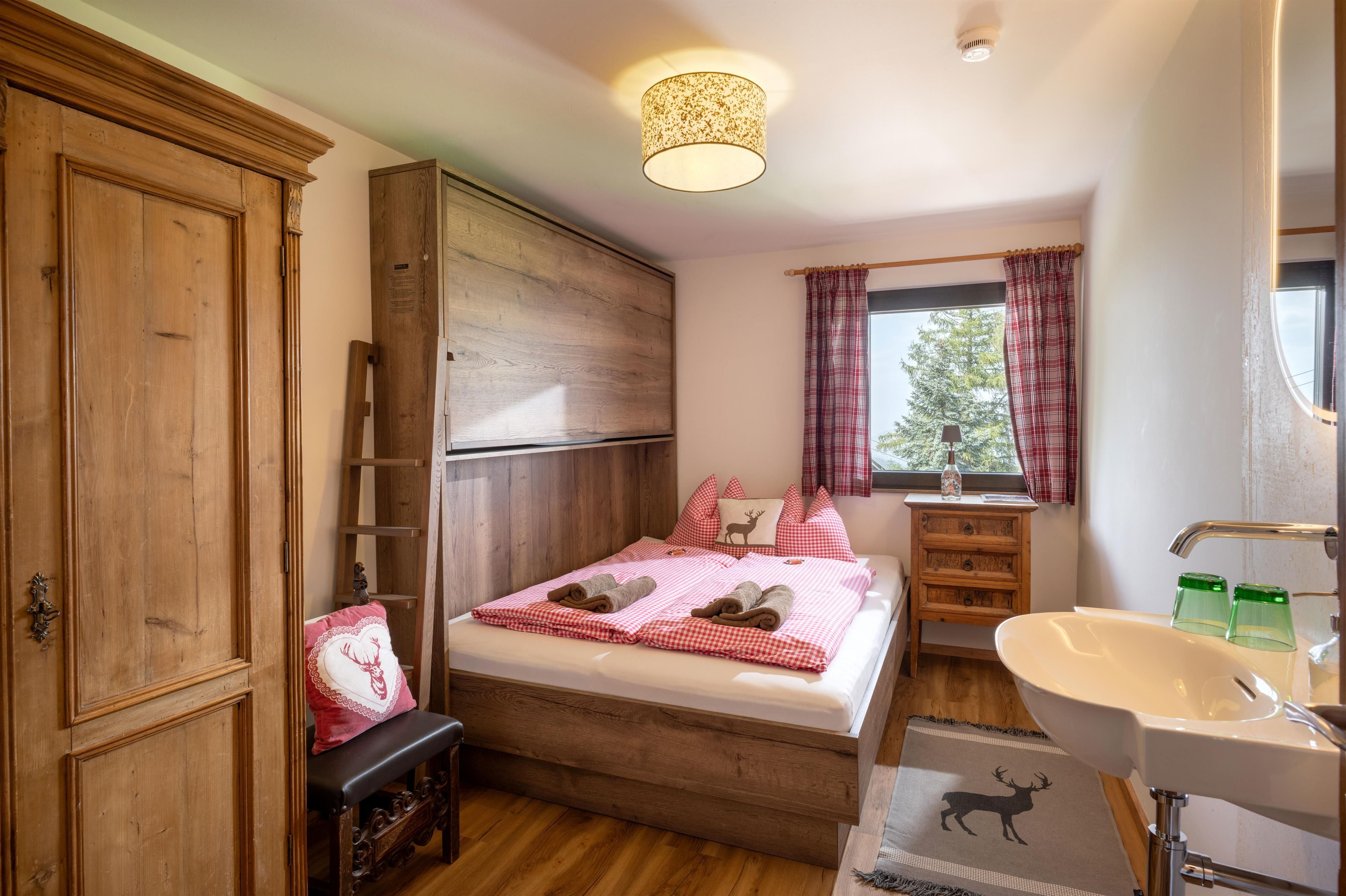 A cozy bedroom with a double bed, wooden wardrobes, and a window. The furnishings are simple, and there is a small washbasin in the room.